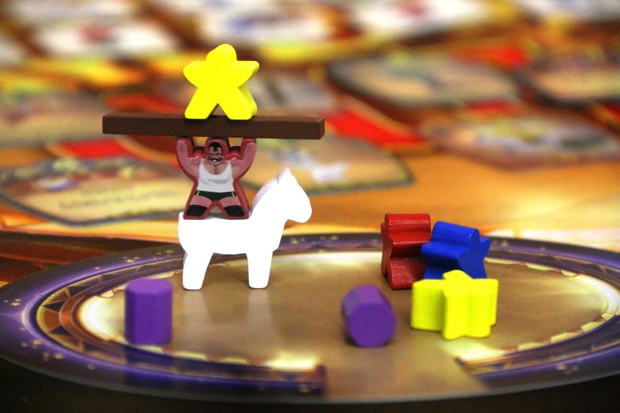 Photo showing gameplay of Meeple CIrcus. Wooden objects are stacked in a cardboad circus ring: a strongman stands on the back of a horse, lifting a plank with another performer on top.